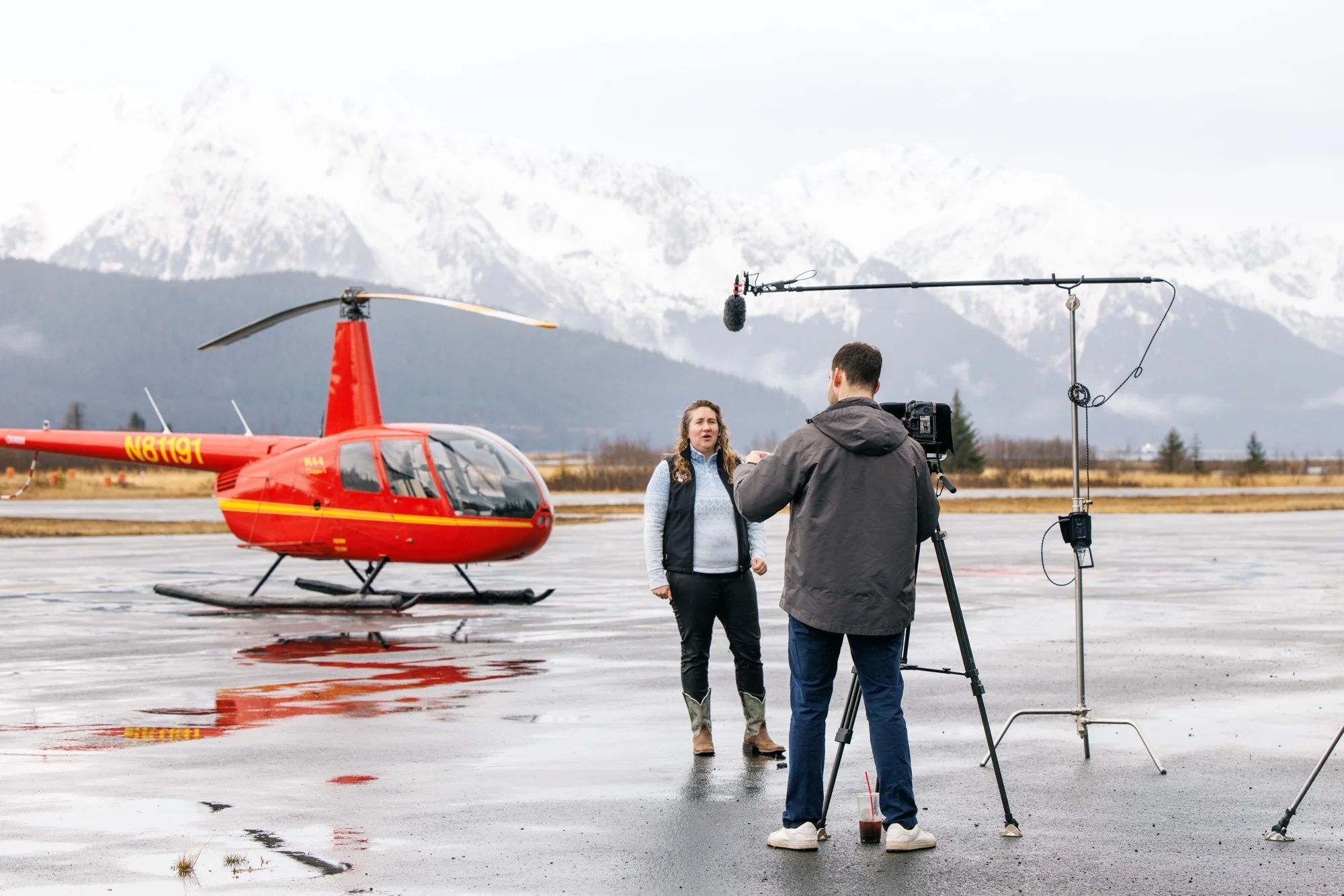 Martin Media filming guide and pilot interview on tarmac in front of red helicopter and snowcapped Chugach mountains